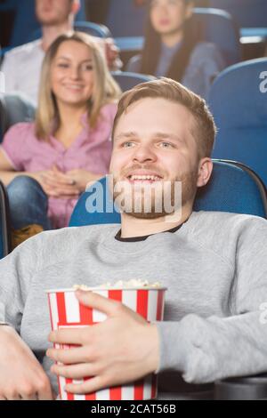 Kinoliebhaber. Vertikale Aufnahme eines hübschen bärtigen jungen Mannes, der Popcornkübel in der Hand hält und sich einen Film im lokalen Kino ansieht Stockfoto