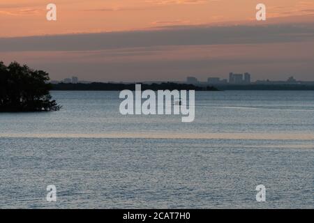 Motorboot geht über einen See mit Bäumen und Gebäuden einer nahe gelegenen Stadt am fernen Horizont in der frühen goldenen Stunde bei Sonnenaufgang. Stockfoto