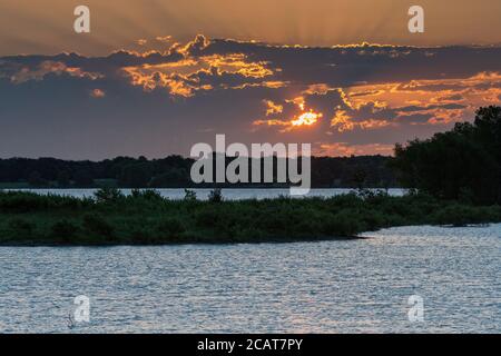 Sonne späht durch die frühen Morgenwolken über einem See mit einer von Bäumen bedeckten Halbinsel, die das Wasser in zwei Seen teilt. Stockfoto