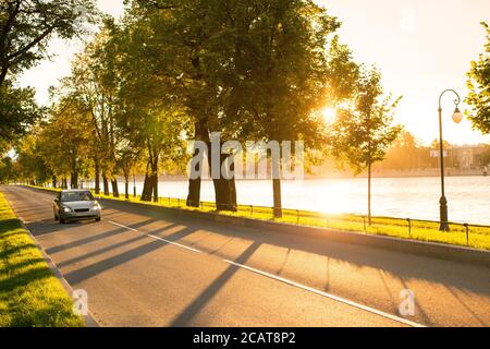 Vorderansicht des modernen Autofahrens oder der Fahrt auf der Asphaltstraße im Park bei schönem Sonnenuntergang, Straßenbeleuchtung und Fluss im Hintergrund. Ziel, automatisch Stockfoto