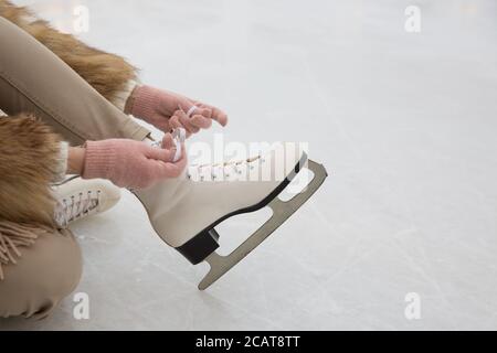 Nahaufnahme des Weibchens in Pelzmantel und rosa Handschuhe tragen Weiße Schlittschuhe auf Eisbahn im Wintertag.Frau binden Schlittschuhe.Wochenenden Aktivitäten im Freien in kalten wir Stockfoto