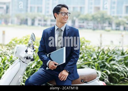 Happy Handsome junge vietnamesische Unternehmer mit Tablet-Computer sitzt auf Roller und wegschauen Stockfoto