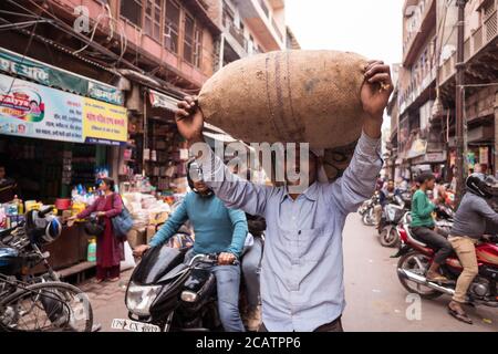 Agra / Indien - 22. Februar 2020: Porträt eines indischen Mannes, der die Straße der historischen Innenstadt von Agra mit schwerem Bündel auf dem Kopf entlang geht Stockfoto