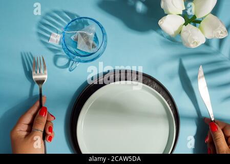 Dinner-Set serviert Keramik-Teller mit Messer und Gabel, Glas von frischem Getränk, Vase mit Blumen auf einem hellblauen Hintergrund mit weichen Schatten aus Stockfoto