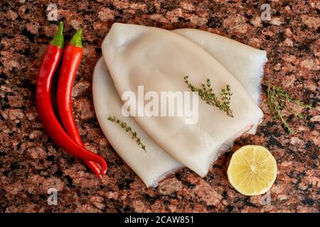 Drei Kadaver von frischem rohem Tintenfisch mit Zitrone, Pfeffer, Thymian auf einem dunklen Marmortisch. Stockfoto
