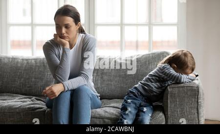 Gestresste junge Mama sitzt getrennt auf der Couch mit beleidigten Sohn. Stockfoto