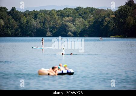 Freiburg, Deutschland. August 2020. Eine Frau reitet auf einem Stand Up Paddle Board (SUP) über den Opfinger See und Badegäste nehmen ein Bad im Wasser. Die sommerlichen Temperaturen ziehen viele Menschen an die Badeseen, aber weniger als befürchtet. Laut DLRG ist die Zahl der bundesweiten Badetodesfälle im Vergleich zum Vorjahr zurückgegangen. Quelle: Philipp von Ditfurth/dpa/Alamy Live News Stockfoto