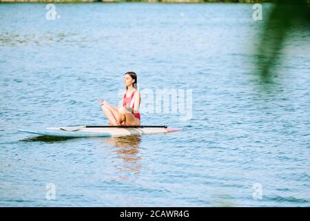 Junge heitere Frau in rotem Badeanzug sitzt auf Surfbrett in Pose des Lotos Stockfoto
