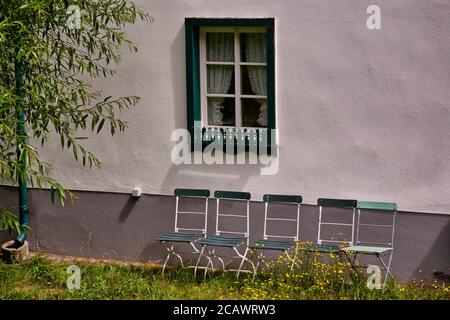 Vier alte grüne verblasste Gartenstühle vor einem Fenster mit Gras und Blumen wachsen um sie herum Stockfoto