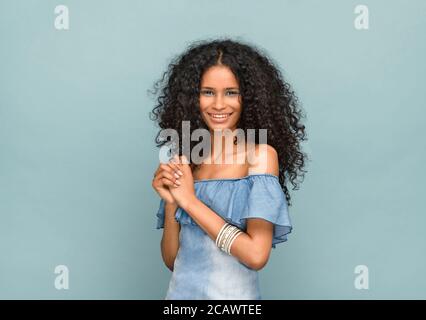 Studio Porträt eines schönen schlanken schwarzen Mädchen mit wunderschönen Lockiges langes Haar, das mit einem strahlenden Blick auf die Kamera schaut Warmes freundliches Lächeln gegen ein blu Stockfoto