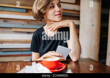 Lächelnde junge Frau, die allein in einem Café sitzt, verloren in einem glücklichen Gedanken Stockfoto