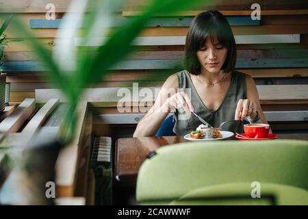 Frau, die gerne Kuchen in einem Café isst. Frontaler Schuss durch eine Pflanze. Stockfoto