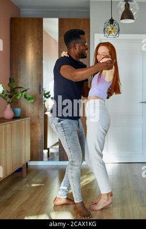 Schöne junge multiethnische Paar tanzen im Wohnzimmer zu Hause, glücklich, in neue Wohnung zu bewegen, schwarzer Mann und Rotschopf Dame sind verliebt Stockfoto