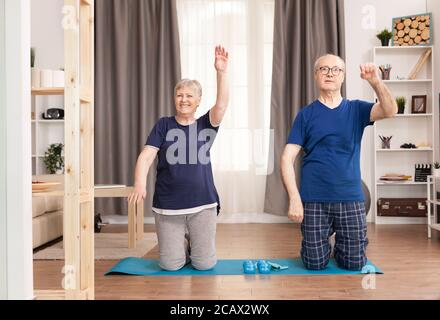 Glückliche alte Frau, die in der Nähe ihres Mannes trainiert. Alter gesunder Lebensstil Bewegung zu Hause, Training und Training, sportliche Aktivität zu Hause auf Yoga-Matte. Stockfoto