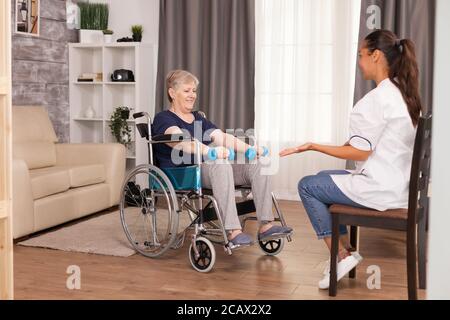 Patient mit Kurzhanteln für die Rehabilitation sitzen im Rollstuhl im Wohnzimmer mit Krankenschwester. Behinderte behinderte alte Person erholt professionelle Hilfe Krankenschwester, Pflege Altersheim Behandlung und Rehabilitation Stockfoto