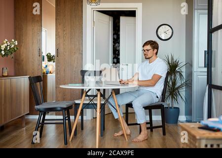 kaukasischen Mann arbeiten auf Laptop zu Hause, junge Mann auf freiberuflichen während Quarantäne. Gut aussehende Männer in Freizeitkleidung sitzen in der Küche Stockfoto