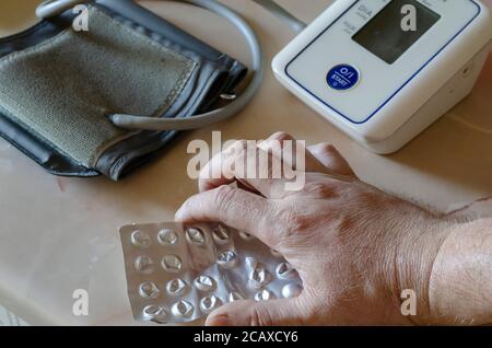 Männliche Hand hält Blister mit Pillen auf dem Tisch. Medizinischer elektronischer automatischer Blutdruckmonitor zur Messung des Blutdrucks im Hintergrund. Trea Stockfoto