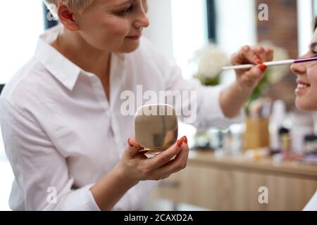 Junge kaukasische Make-up-Künstler gehen, um Pulver auf das Gesicht des Modells gelten, verwendet sie professionelle dekorative Kosmetik Suring Arbeit Stockfoto