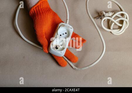 Die Hand trägt einen orangefarbenen Schutzhandschuh und hält nach dem Brand eine Steckdose. Weiße Steckdosenleiste nach Kurzschluss. Elektrische Sicherheit. Stockfoto