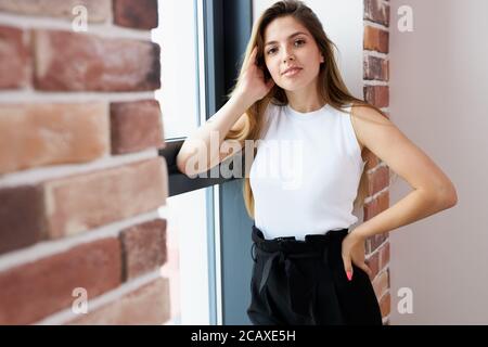 Porträt der jungen attraktiven kaukasischen Frau posiert vor der Kamera neben dem Fenster, sie schaut auf Kamera und lächeln. Schöne Frau mit langen Haaren steht ne Stockfoto