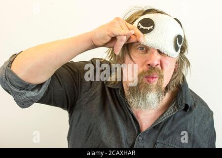 Mann mit Bart zieht seine Schlafmaske aus. Verärgert müde Mann hat nicht genug Schlaf, berührt Gesicht mit der Hand, trägt Augenmask und, schlief schlecht in der Nacht. Stockfoto