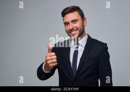 Geschäftsmann Geschäftsmann. Mann Business Anzug Studio grau Hintergrund. Moderne Millennial Person zeigt Daumen nach oben Zeichen. Porträt von charmant erfolgreich Stockfoto