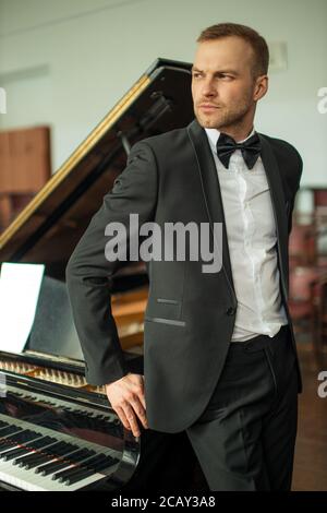 Porträt eines schönen kaukasischen Pianisten in elegantem Anzug mit Fliege posiert vor der Kamera und Blick Seite, neben klassischen Instrument Klavier Stockfoto