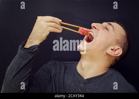 Mann isst Brötchen mit Essstäbchen. Öffnen Sie den Mund. Schwarzer Hintergrund Stockfoto