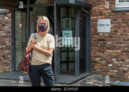 Eine Frau mit einer gemusterten Gesichtsmaske verlässt das Auerbach-Haus auf der Stadtparkinsel in Grevenbroich, Nordrhein-Westfalen, Deutschland, nach einem Coronatest; ein Schild am Eingang identifiziert das Gebäude als „Corona-Test-Zentrum“ (Coronatestzentrum), ein zweites nennt es „Auerbach – Haus“. Die Fotografie wurde im Juli 2020 während der Coronavirus-Pandemie aufgenommen. Das Backsteingebäude, das sonst als Schulungsgebäude des örtlichen Volkshochschulzentrums dient und nach der Partnerstadt Auerbach benannt ist, diente vorübergehend als Coronatestzentrum des Rhein-Kreises Neuss während der Pan Stockfoto