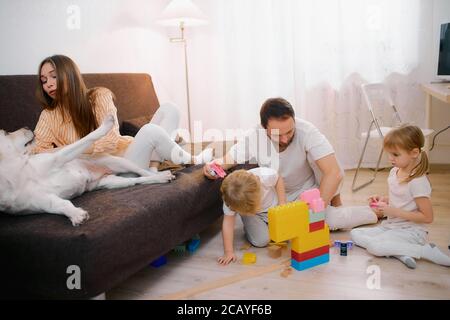 Junge bärtige Vater spielen mit Kindern auf dem Boden, schöne Mutter streichelt freundlich weißen Hund auf dem Sofa sitzen, jeder in weißen Casual Domest Stockfoto