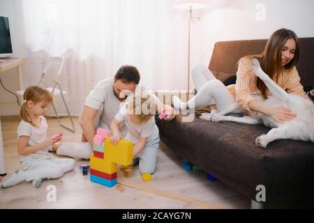 Freundliche kaukasische Familie haben verspielte Haustier, weißen Hund zu Hause. Mann, Frau und Kinder im Wohnzimmer, spielen mit Kindern und mit Hund, tragen lässig Stockfoto