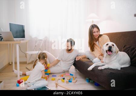 Junge bärtige Vater spielen mit Kindern auf dem Boden, schöne Mutter streichelt freundlich weißen Hund auf dem Sofa sitzen, jeder in weißen Casual Domest Stockfoto