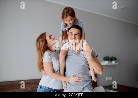 Fröhliche junge kaukasische Familie, Eltern mit Tochter, posiert bei der Kamera isoliert im Schlafzimmer, umarmt sich gegenseitig Stockfoto