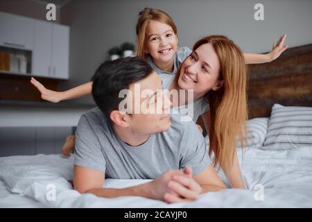 Schöne kaukasische Eltern liegen auf dem Bett mit ihrem Kind Mädchen, tragen lässige Kleidung, glücklich lächelnde Familie zusammen zu Hause Stockfoto