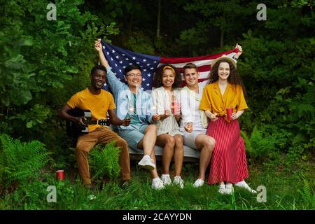 In voller Länge Porträt der multi-ethnischen Gruppe von Menschen halten amerikanische Flagge, während auf der Bank im Wald sitzen und genießen Sommerferien, kopieren Raum Stockfoto