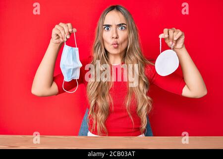 Junge schöne blonde Frau hält zwei verschiedene Sicherheitsmasken machen Fisch Gesicht mit Mund und Augen schielen, verrückt und komisch. Stockfoto