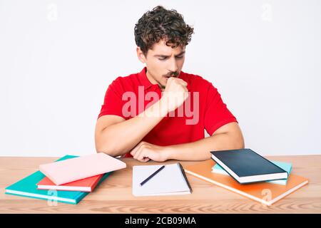 Junge kaukasische Mann mit lockigen Haaren auf dem Tisch sitzen stuying für Universität Gefühl unwohl und Husten als Symptom für Kälte oder Bronchitis. Gesundheit Stockfoto