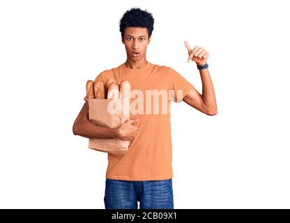 Junger afroamerikanischer Mann, der Papiertüte mit Brot hält, das mit Fingern nach unten zeigt und Werbung zeigt, überrascht das Gesicht und den offenen Mund Stockfoto