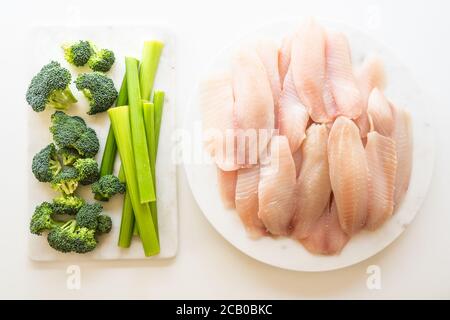 Weißes Fischfilet und frisches Gemüse. Rohes Tilapia-Fischfilet, Brokkoli und Sellerie aus der Nähe auf weißem Hintergrund, Ansicht von oben Stockfoto