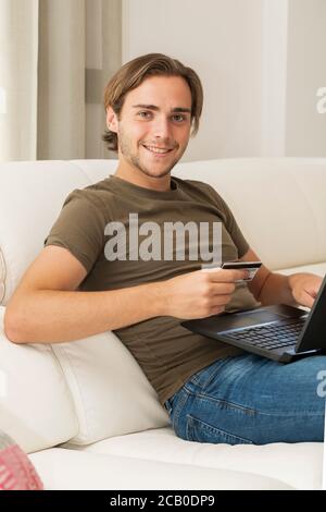 Lächelnder, hübscher junger Mann, der auf einem Sofa sitzt, während er einen Laptop und eine Kreditkarte auf einem unfokussieren Hintergrund hält. Einkaufs- und Zahlungskonzept. Stockfoto