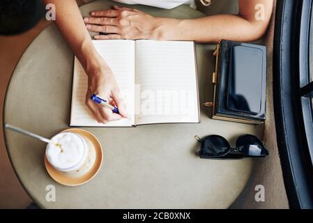 Hände der jungen Frau trinken Cappuccino und schriftlich Pläne für den Tag in Planer, Blick von oben Stockfoto