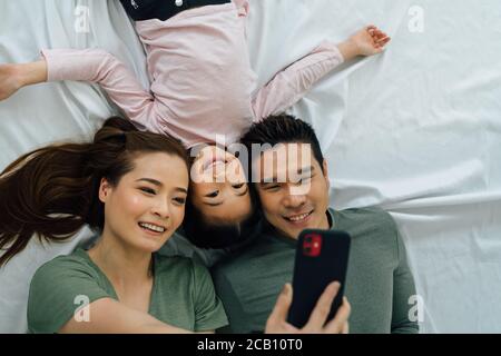 Asiatische Mutter und Vater liegen mit niedlichen Tochter auf dem Bett fotografieren mit Smartphone. Familienbindung und Glück Lifestyle-Konzept. Stockfoto