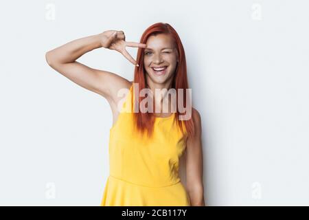 Glückliche Dame mit roten Haaren lächelt auf einem weißen Studio Wand, die das Friedenszeichen mit zwei Fingern gestikuliert Stockfoto