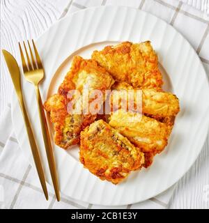 Gebratenes Seehechtfischfilet, serviert auf einem weißen Teller mit goldenem Besteck auf weißem Holzhintergrund, Draufsicht, flaches Lay, Nahaufnahme Stockfoto