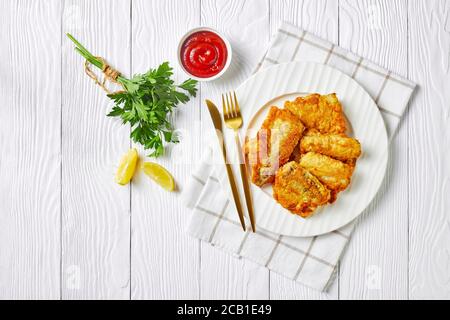 Gebuttertes Kabeljaufilet im britischen Stil, serviert auf einem weißen Teller mit Besteck, Tomatenketchup, frische Petersilie auf weißem Holzhintergrund, Top Vie Stockfoto