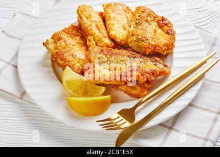 Eine Portion gebratenes Seehechtfischfilet, serviert auf einem weißen Teller mit Zitronenkeilen goldenes Besteck auf weißem Holzgrund mit Serviette, Stockfoto