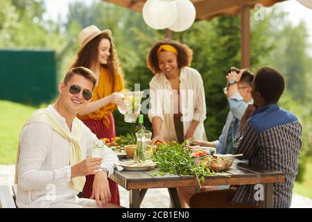 Multiethnische Gruppe von Freunden, die im Sommer auf der Außenterrasse speisen, konzentrieren sich auf junge Männer, die Sonnenbrillen tragen und die Kamera anlächeln, Platz kopieren Stockfoto