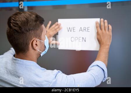 Eröffnung des Geschäfts nach der Sperrung. Rückansicht eines jungen Mannes in Schutzmaske aufkleben offen Schild mit Text SIND WIR AUF der Glastür GEÖFFNET Stockfoto