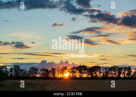 Magischer Sonnenuntergang mit Sonne tief über den Bäumen und dramatischen Wolken beleuchtet von letzten Sonnenstrahlen. Toller Sommerabend Stockfoto