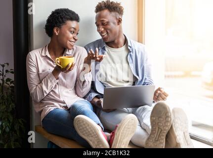 Fröhliches afroamerikanisches Paar mit Laptop und Kaffee Fensterbank im Café Stockfoto